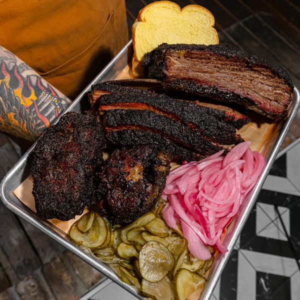 A tray of Sanders BBQ brisket, ribs, and chopped meat with pickled onions, pickles, and bread.