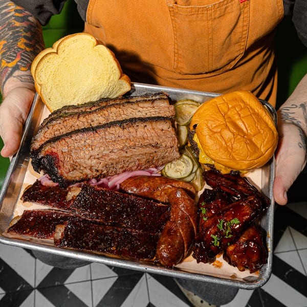 Hands holding a metal tray with Sanders BBQ brisket, ribs, sausage, wings, pickles, and bread.