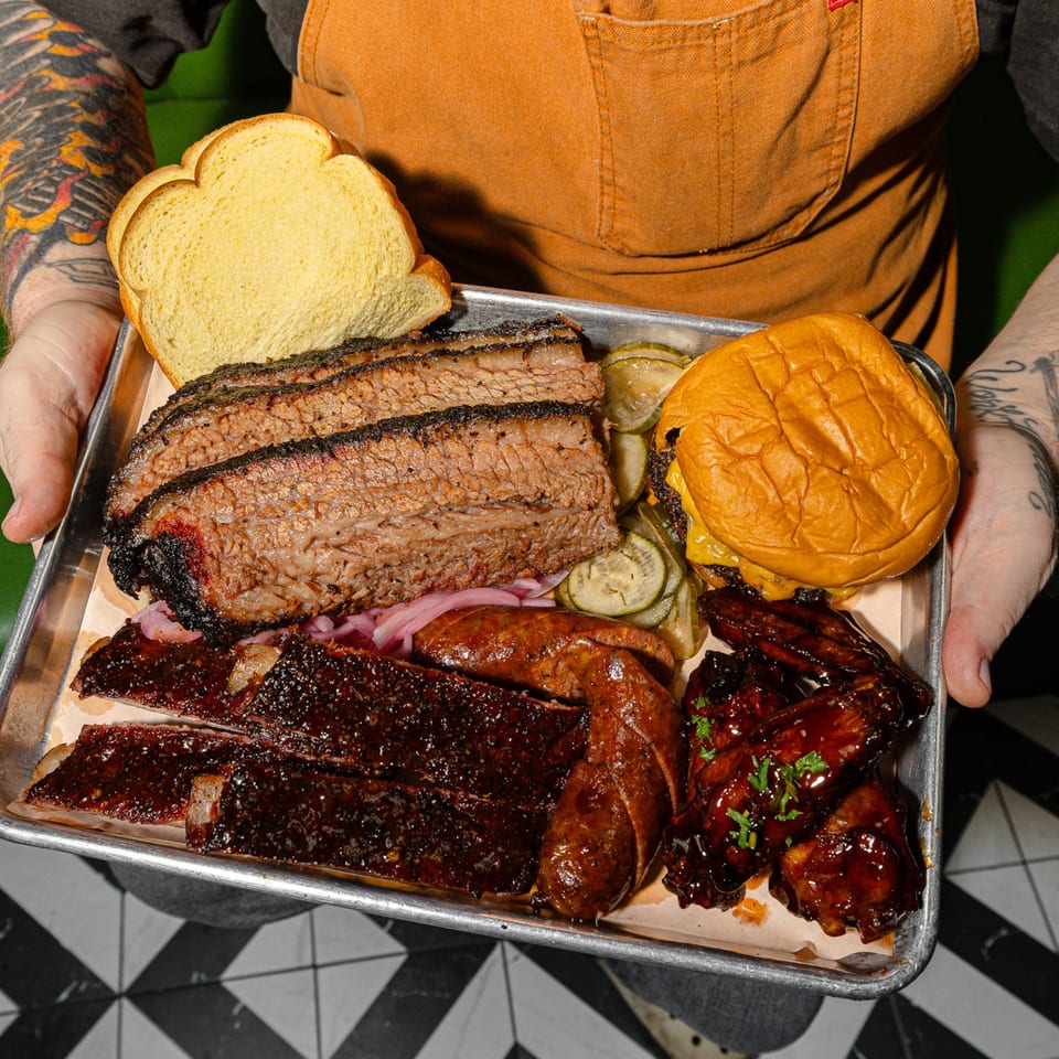 Hands holding a metal tray with Sanders BBQ brisket, ribs, sausage, wings, pickles, and bread.