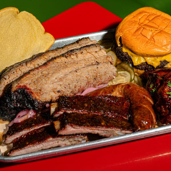 A metal tray filled with Sanders BBQ brisket, ribs, sausage, a cheeseburger, and pickles.