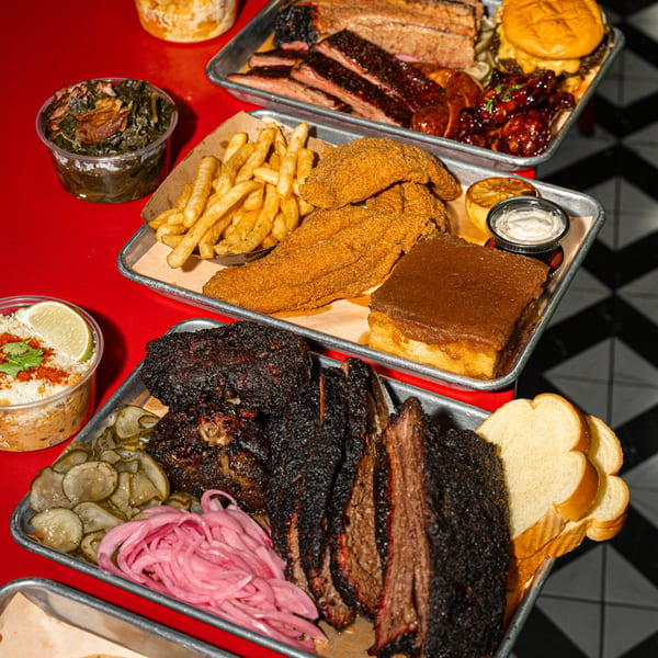 Multiple trays of Sanders BBQ, including brisket, ribs, fried fish, fries, and sides on a red table.